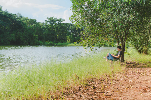relajarse y la actividad de estilo de vida de hombre asiático de 30 a 40 años durante sentarse y leer libro al lado del río y bajo el árbol en la temporada de primavera con sensación de felicidad con fondo de la puesta del sol
 - Foto, Imagen