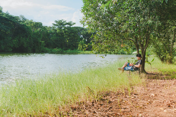 ontspannen en levensstijl activiteit van Aziatische man 30s tot 40s tijdens zitten en lezen boek aan de zijkant van de rivier en onder de boom in het voorjaar met geluk gevoel met zonsondergang achtergrond - Foto, afbeelding
