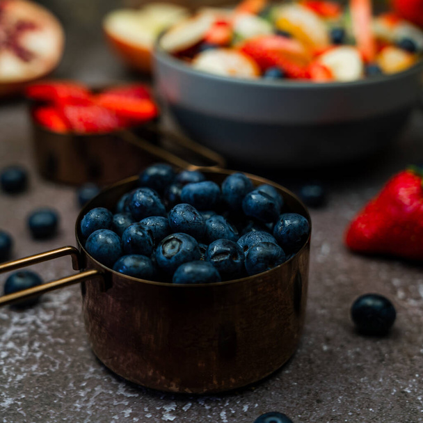 close up view heaps of blueberries in copper measuring cup with fruit salad bowl and strawberries in the background - 写真・画像