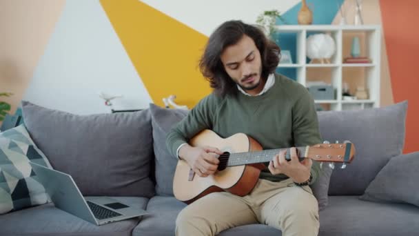 Creative Arab man playing the guitar and using laptop watching tutorial at home - Кадры, видео