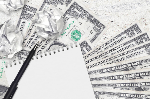 1 δολάριο ΗΠΑ χαρτονομίσματα και μπάλες από τσαλακωμένο χαρτί με σημειωματάριο. Κακές ιδέες ή λιγότερο της έννοιας της έμπνευσης. Αναζήτηση ιδεών για επενδύσεις - Φωτογραφία, εικόνα