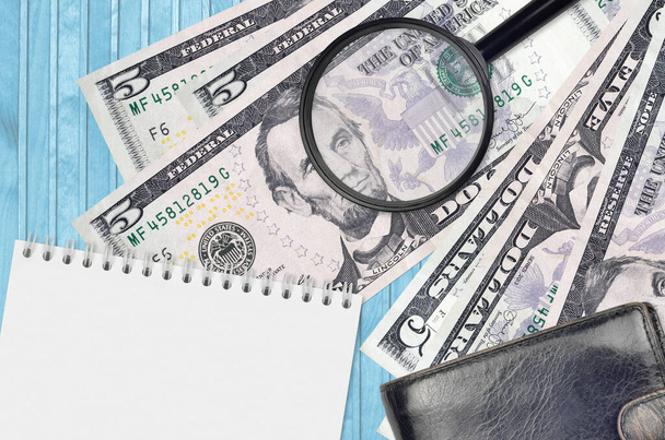 5 US dollars bills and magnifying glass with black purse and notepad. Concept of counterfeit money. Search for differences in details on money bills to detect fake money - Photo, Image