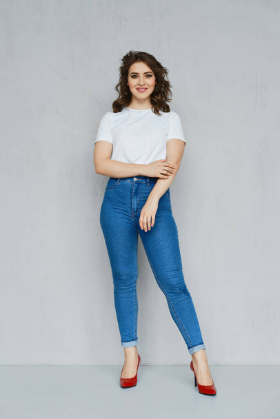 Positive girl in jeans, white t-shirt and red stilettos posing near the wall holding hand down - Fotoğraf, Görsel