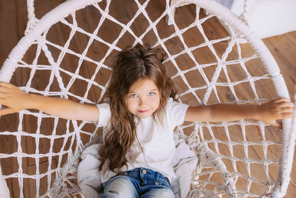 Schattig vrolijk klein meisje in wit t-shirt en jeans in stijlvolle schommelstoel indoor. Mode, stijl, kindertijd, emoties, opgroeiconcept - Foto, afbeelding