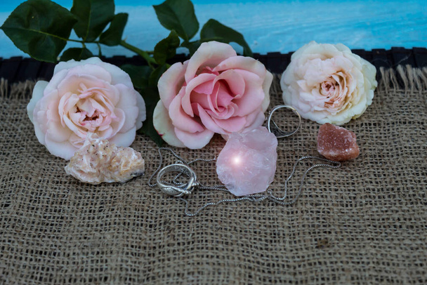 На джуте ткани полудрагоценные камни розовый кварц, доломит, галит. Рядом серебряные кольца и цепь. На заднем плане три розовых розы
 - Фото, изображение