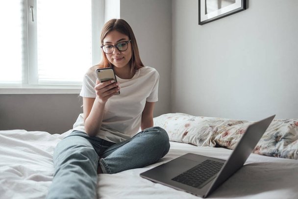 Young smiling woman use smartphone at home on the bed. Communication, video call, remote work. High quality photo - Foto, Imagem
