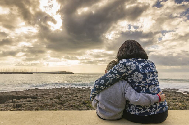 Mãe com o filho sentado e abraçando uns aos outros na parede de frente para o mar
 - Foto, Imagem