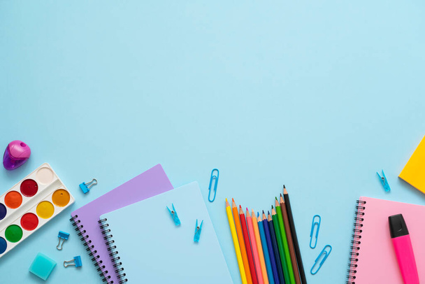 Bright colourful school stationery on blue background. Top view of blue, purple and pink notebooks, color pencils, watercolor, paper clips, marker and eraser. Copy space. - Photo, image