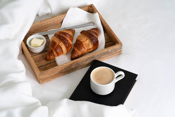 Breakfast in bed with two croissants and butter on wooden tray, cup of coffee and book on white sheet with blanket. Time for yourself.  - 写真・画像