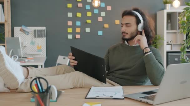 Happy office worker Arab guy relaxing in workplace enjoying music in headphones using laptop - Πλάνα, βίντεο