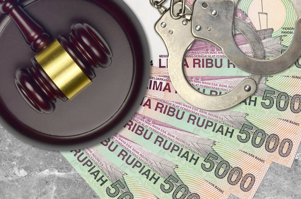 5000 Indonesian rupiah bills and judge hammer with police handcuffs on court desk. Concept of judicial trial or bribery. Tax avoidance or tax evasion - Photo, Image