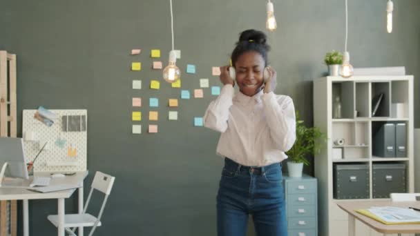 Cheerful mixed race business lady wearing headphones dancing in office room - Metraje, vídeo