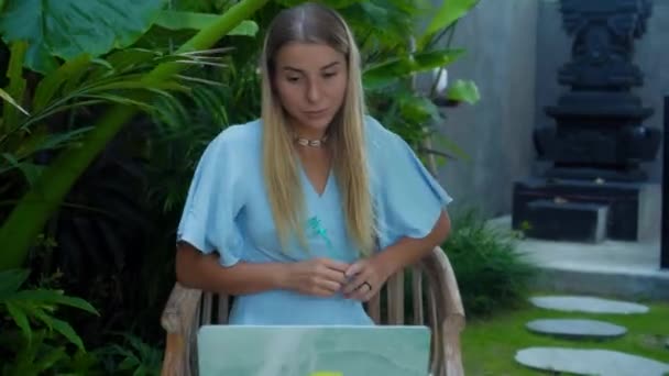Video of a young girl sitting at a wooden table and working behind a laptop in a green garden - Materiał filmowy, wideo