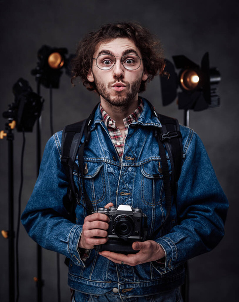 Joven hombre caucásico en gafas con chaqueta de mezclilla sostiene una cámara digital, se ve sorprendido
 - Foto, imagen