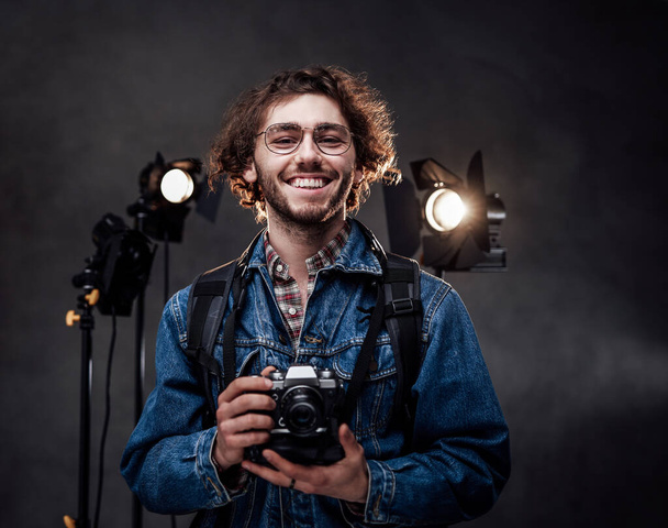 Gözlüklü, kot ceket giyen mutlu, kıvırcık bir adamın portresi karanlık fotoğraf stüdyosunda bir dijital kamera tutuyor. - Fotoğraf, Görsel