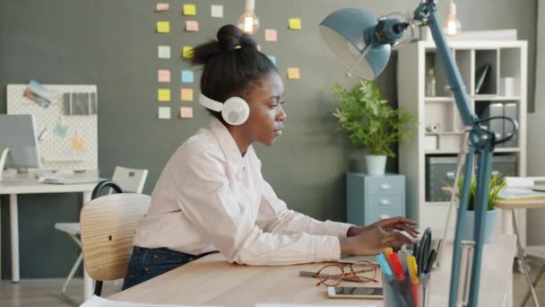 Slow motion of joyful mixed race girl working with laptop then dancing wearing headphones in office - Video