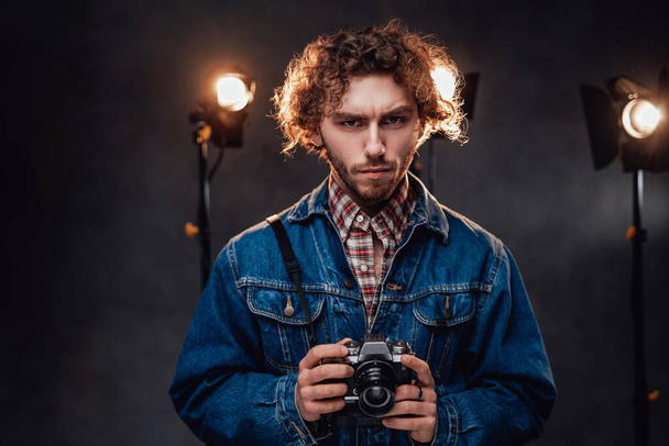 Schöner Mann mit lockigem Haar hält eine Digitalkamera in der Hand und schaut in die Kamera, sieht ernst aus - Foto, Bild