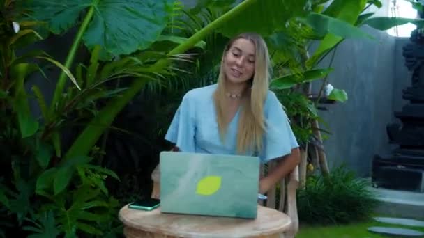Video of a young girl sitting at a wooden table and working behind a laptop in a green garden - Materiaali, video
