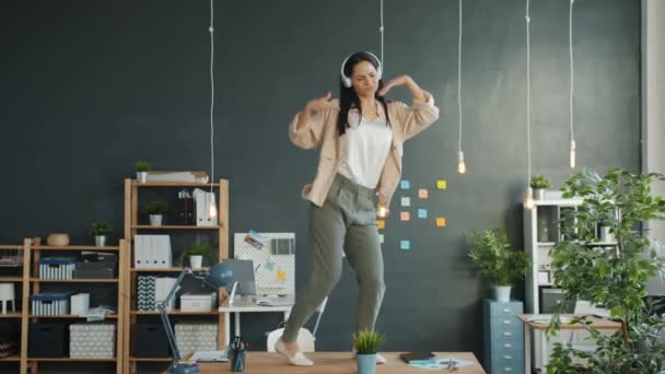 Happy businesswoman dancing on office desk fooling around enjoying music in headphones - Felvétel, videó