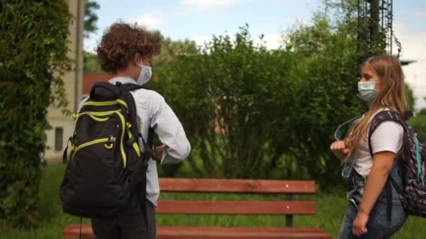 Masked students greet each other contactless. Back to school after quarantine. Boy and girl in protective masks sit on a bench at a safe distance, social distance - Záběry, video