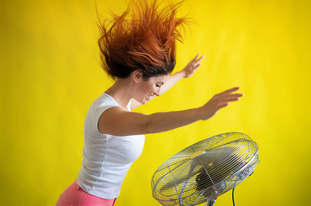 A beautiful red-haired woman is cooled off standing over a large electric fan on a yellow background. Girl with hair developing in the wind. Device for cooling the air. - Photo, image