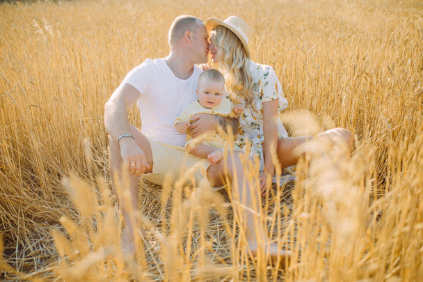 Happy young parents with baby rests among yellow wheat field and they kisses. - Photo, Image