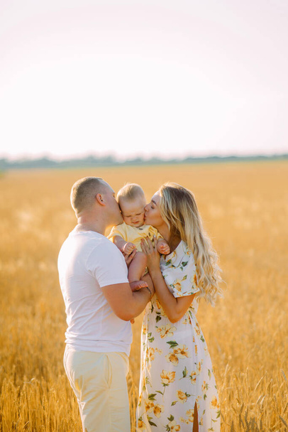 Happy young parents walk with their baby and kiss his among yellow wheat field. - Фото, зображення