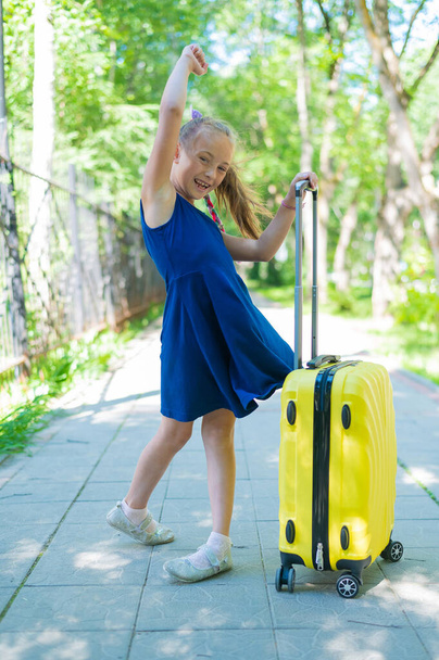 A smiling little girl in a blue dress holds a yellow suitcase and dances from happiness outdoors. The child is ready for a summer vacation trip. - Фото, изображение