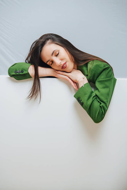 Happy gorgeous sensual young woman resting on an edge of an empty bathtub, having sweet nap, her eyes closed. She's wearing green jacket. - Foto, Imagem