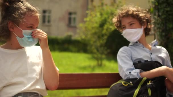 School-age children in medical masks sit on a bench and then say goodbye, touching each other with their elbows. Contactless farewell, back to school after quarantine coronavirus covid-19 - Πλάνα, βίντεο