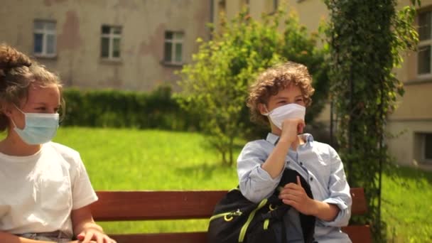 A boy and a girl, masked schoolchildren are sitting on a bench in the school yard. A classmate comes up and greets touching her elbow with each. Contactless welcome, new rules - Video, Çekim