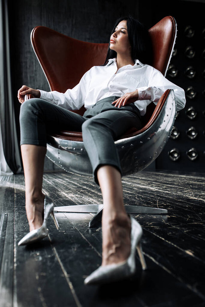 Business shot of a business girl who is sitting in a suit in an aviator armchair and is scattering papers in a loft room. - 写真・画像