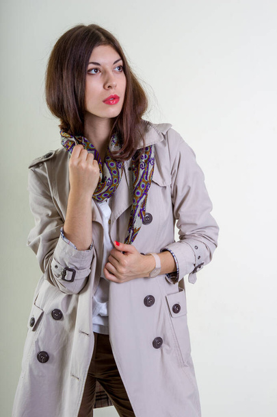 A beautiful fashionable woman in a light raincoat and a neckerchief poses on a light background. - Foto, Bild