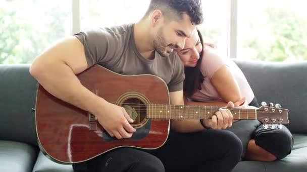 Мужчина играет на гитаре со своей женой подружкой дома в гостиной. Мужчина играет любовные песни для любовника. Концепция празднования, счастливая пара играет на гитаре и поет песню вместе в гостиной
. - Кадры, видео