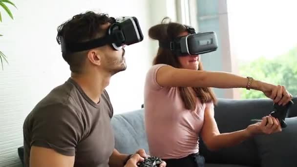 Couple wearing VR or virtual reality glasses, headsets sitting and playing a video game on sofa at home in technology and innovation concept. Having fun. Entertainment concept. - Filmmaterial, Video