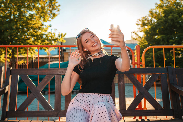 Egy boldog nő ül egy padon egy parkban, és kommunikál videó linken keresztül egy okostelefonon. Nyár. A társadalmi függőség és a távközlés fogalma. - Fotó, kép