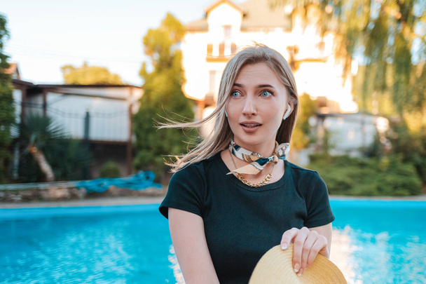 An elegant woman looks away in surprise and fear, holding her hat in her hands. In the background there is a pool and a house. Concept of human emotions and expressions. - Photo, Image