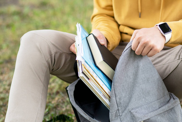 Student, pupil boy puts textbooks, books in a backpack outdoors sitting on grass in a park - Photo, image