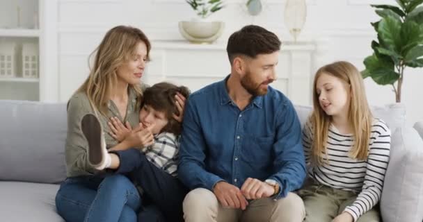 Portret wesołej białej rodziny z dwójką dzieci siedzących na kanapie i uśmiechających się radośnie podczas przytulania i rozmowy. Mały słodki chłopiec i dziewczyna z matką i ojcem w domu. Rodzice z małymi dziećmi - Materiał filmowy, wideo