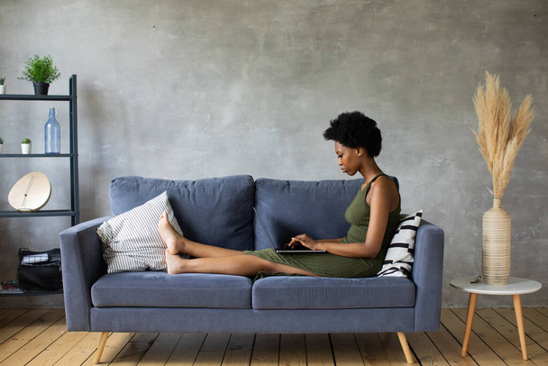 Ευτυχισμένη Αφροαμερικανή νεαρή γυναίκα κάθεται χαλαρώστε σε άνετο καναπέ και εργάζονται σε ένα φορητό υπολογιστή. Χαρά μου να μετακομίσω σε νέο διαμέρισμα - Φωτογραφία, εικόνα
