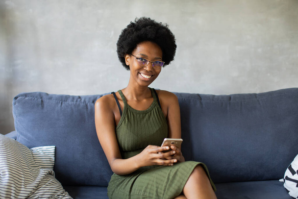 Happy african American young woman looking in camera sit relax on cozy couch happy to move to new apartment. Smiling black girl rest on comfortable sofa in living room dreaming - Photo, Image