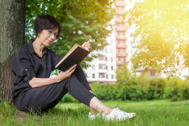 Beautiful elderly mature adult woman sitting on the ground, grass in the summer park, reading an interesting book in glasses, leaning on a tree in a sunny day - Фото, зображення