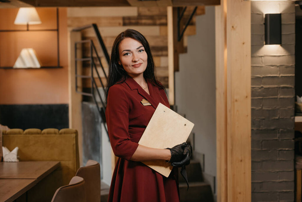 A smiling female restaurant manager in black disposable medical gloves is posing holding a wooden menu in a restaurant. A kind cafe owner in a ruby dress. - Foto, Bild