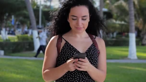 Young woman or girl is texting messages on the phone on a sunny day on a background of palm trees, greenery, trees on a city park. - Video
