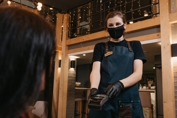 Μια σερβιτόρα που φοράει μάσκα προσώπου και γάντια μιας χρήσης παραδίδει ένα ασύρματο τερματικό πληρωμής σε μια πελάτισσα σε ένα εστιατόριο. Το προσωπικό περιμένει τον καταναλωτή να πληρώσει ένα λογαριασμό.. - Φωτογραφία, εικόνα