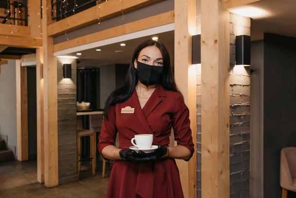 Μια πανέμορφη γυναίκα διευθύντρια εστιατορίου που φοράει μαύρη μάσκα προσώπου και γάντια μιας χρήσης κρατάει ένα φλιτζάνι καφέ σε ένα εστιατόριο. Ένας ευγενικός καφετζής με ρουμπινί φόρεμα περιμένει τους πελάτες.. - Φωτογραφία, εικόνα