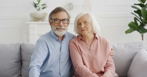 Portret van blanke laatstejaars gepensioneerden die thuis op de bank zitten. Oude man en vrouw kijken naar de camera op de bank in knuffels in de woonkamer. Man en vrouw bij pensionering. - Video