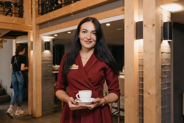 Eine wunderschöne Restaurantmanagerin hält in einem Restaurant eine Tasse Kaffee in der Hand. Eine freundliche Cafébesitzerin in einem rubinroten Kleid wartet auf eine Kundin. - Foto, Bild