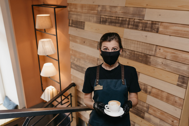 Een mooie serveerster die een zwart gezichtsmasker draagt en wegwerphandschoenen houdt een kop koffie vast in een restaurant. Een schattige barista wacht op klanten in een café. - Foto, afbeelding