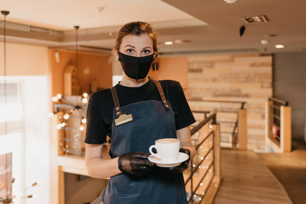 A waitress who wears a black face mask and disposable gloves is serving a cup of coffee on the second floor of a restaurant. A cute barista is waiting for clients in a cafe. - Фото, изображение